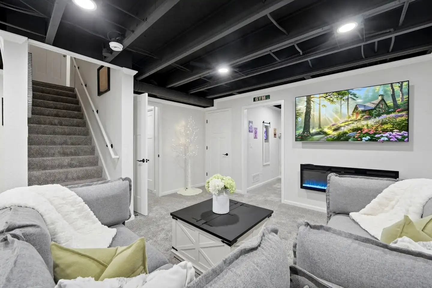A modern basement remodel featuring a sleek built-in entertainment wall with an electric fireplace, large TV, and rustic live-edge floating shelves on both sides. A sliding barn door and light hardwood floors complete this clean, functional Philadelphia basement finishing project.